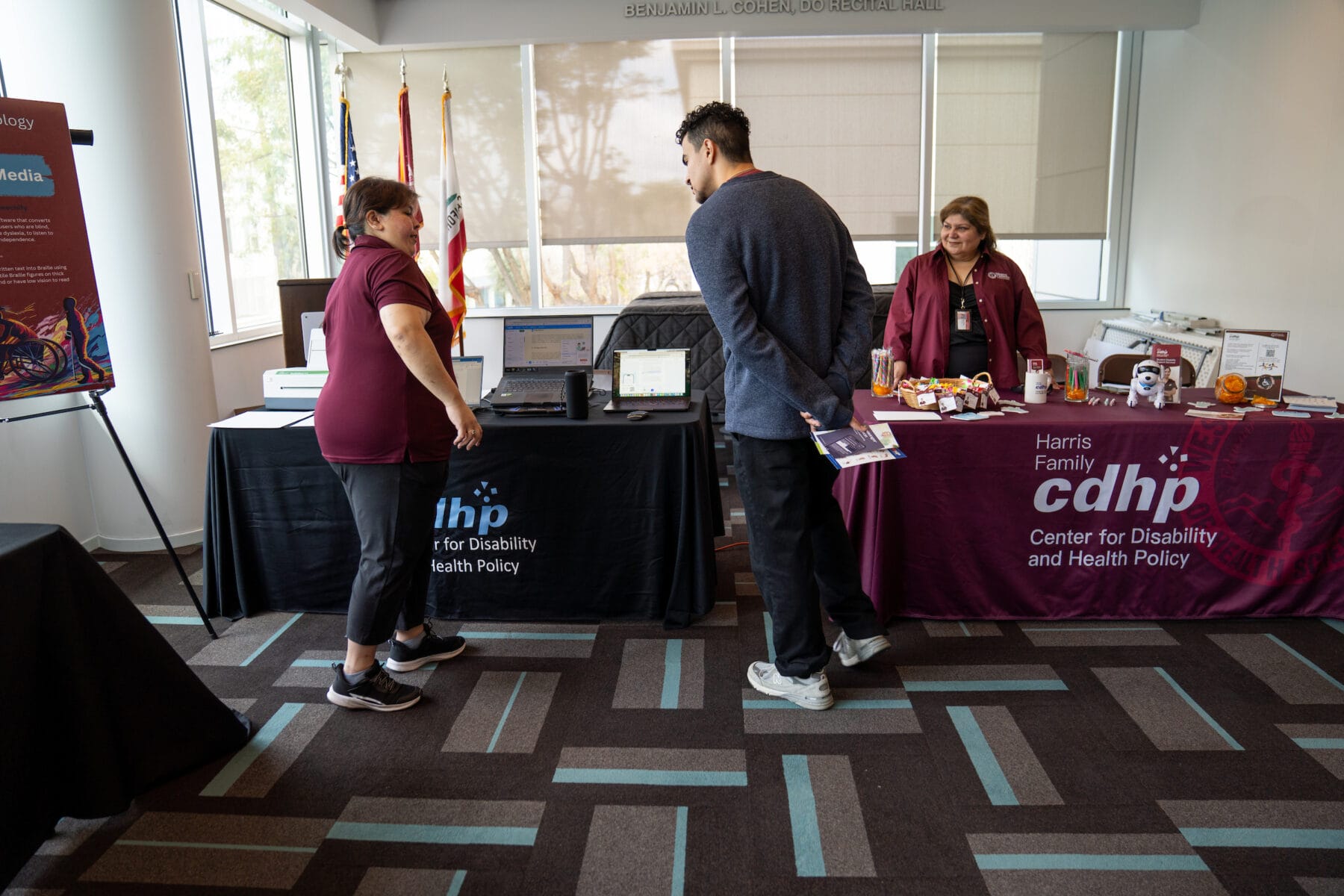 Two women stand behind tables with informational displays for the Center for Disability and Health Policy while a man speaks with them at an indoor event.