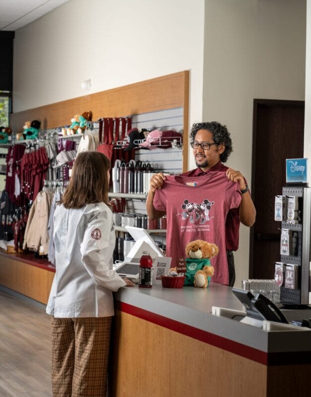 A man behind a store counter holds up a maroon t-shirt with a dog graphic for a woman in a white coat; merchandise and plush toys are displayed on shelves.