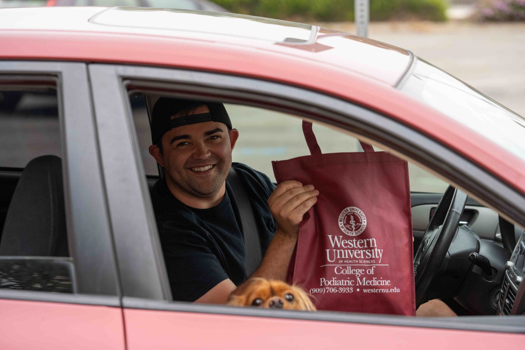 Man in a red car holding a Western University College of Podiatric Medicine tote bag, with a small brown dog visible at the window.