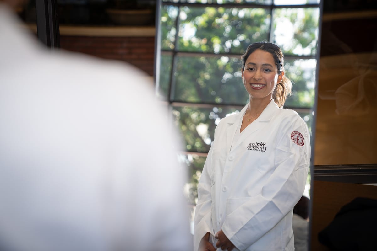 A woman in a white lab coat stands indoors, smiling at her reflection in a mirror with a window and greenery visible behind her.