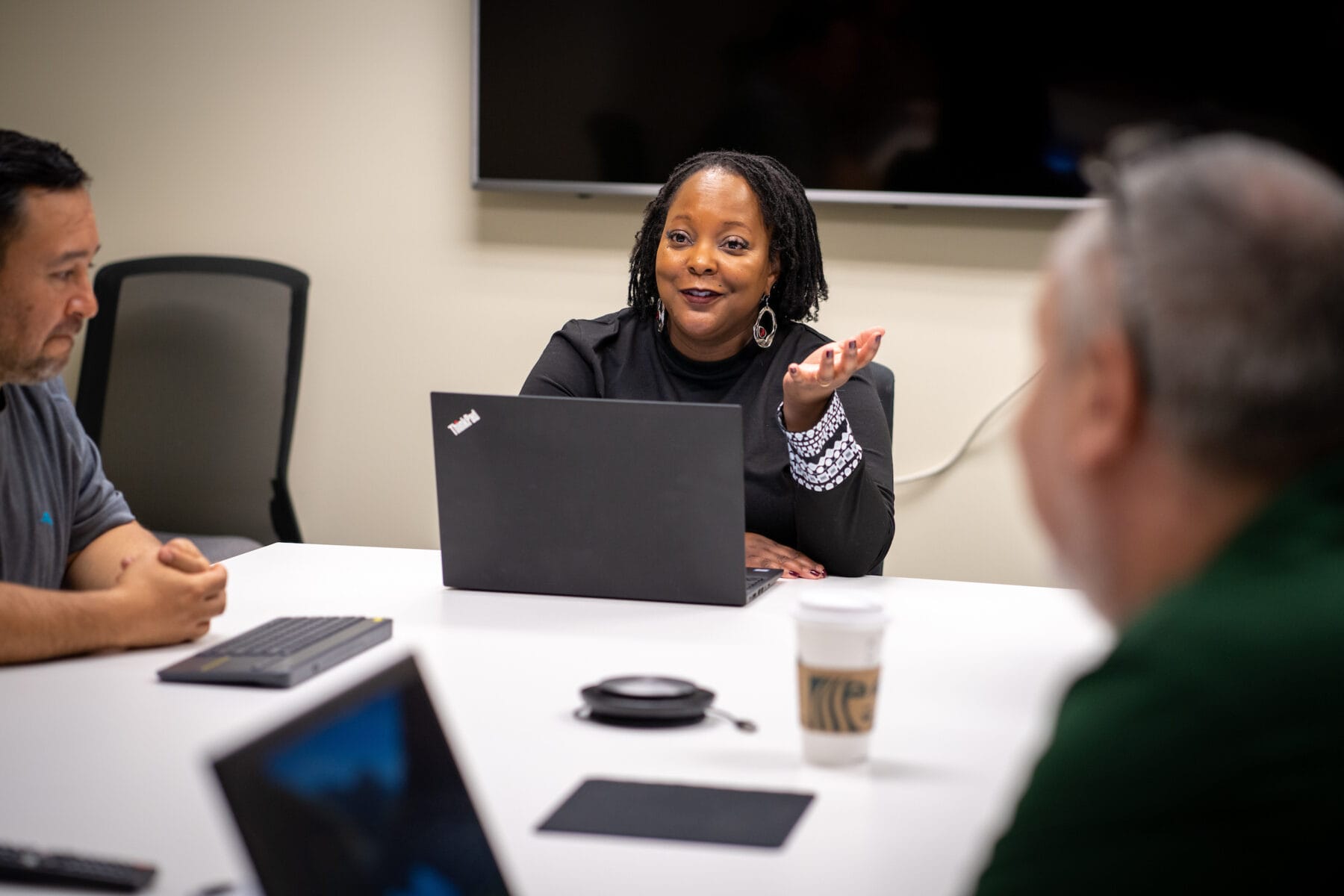 A woman speaks during a meeting, seated at a table with a laptop and coffee cup, while two other people listen.