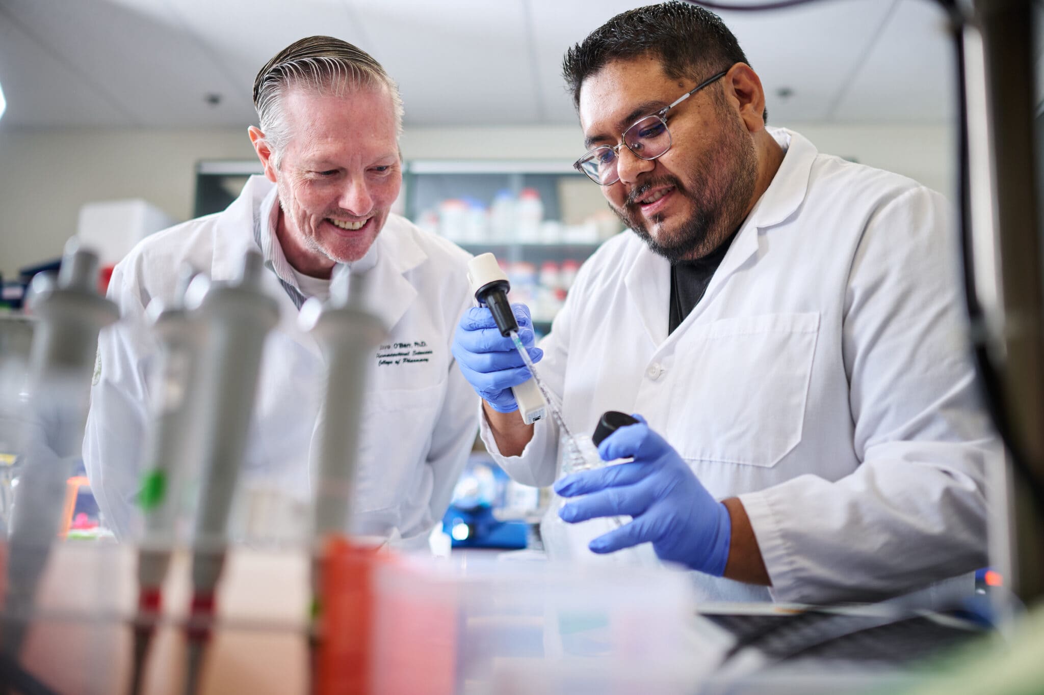 Two scientists in lab coats and gloves working with a pipette and a vial in a laboratory.