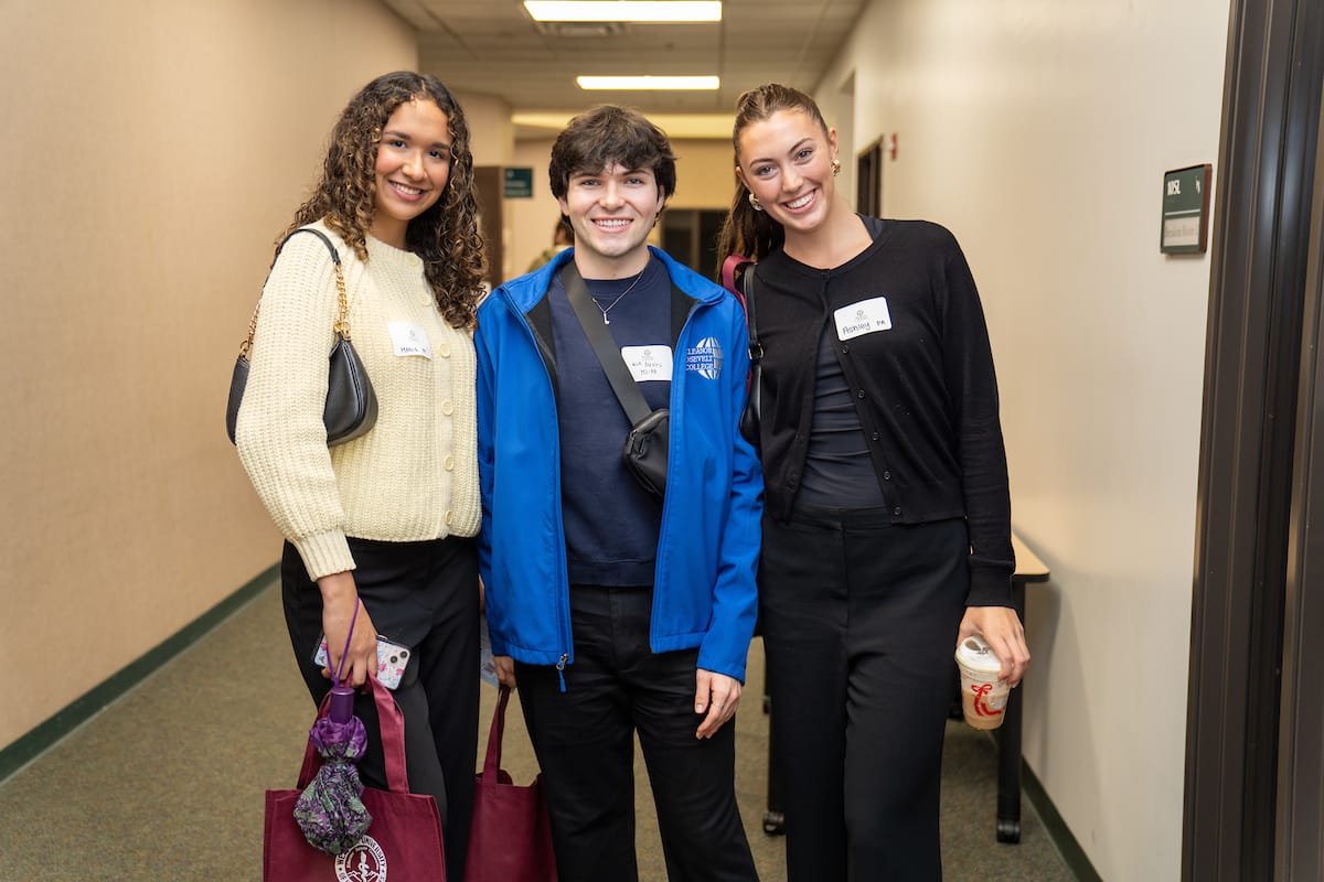 Three people stand in a hallway, smiling at the camera. They wear name tags and carry bags; one holds a beverage cup. The setting appears to be indoors in an institutional building.