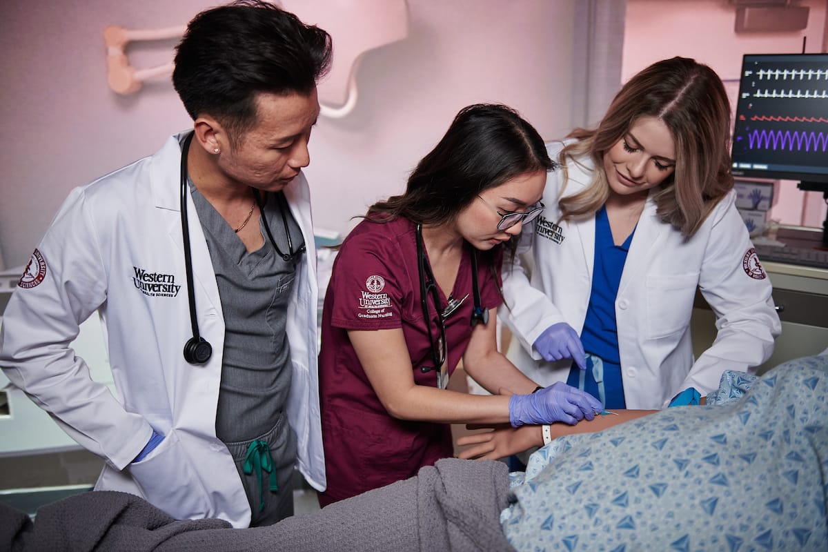 Three medical professionals stand by a hospital bed, one inserting an IV into a patient’s arm while the others observe. Medical monitors and equipment are visible in the background.