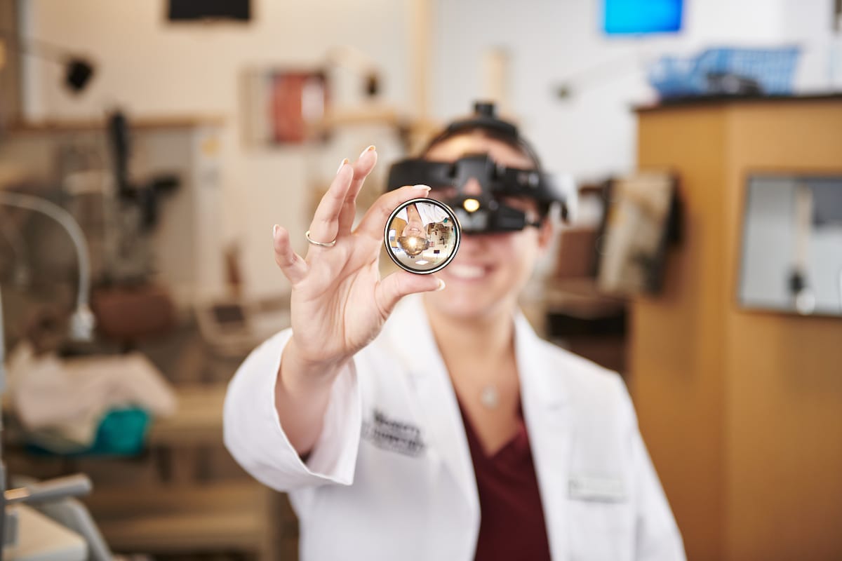 Optometrist holds a lens in front of the camera, wearing specialized eye examination equipment in a clinical setting.