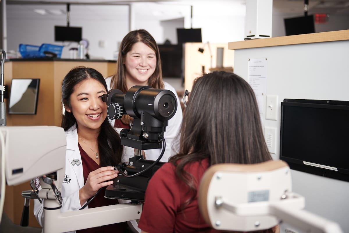 Two women in medical attire observe as a third woman uses an ophthalmic device for an eye examination in a clinical setting.