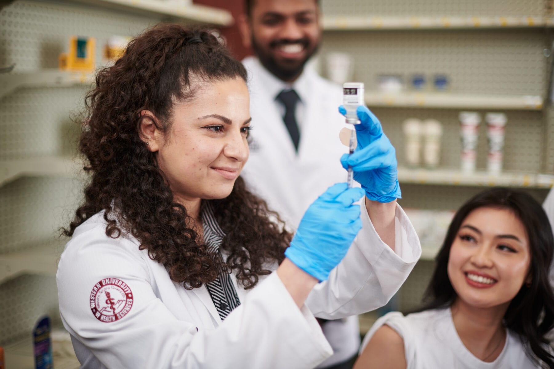 A woman in a lab coat and blue gloves draws liquid from a vial with a syringe, while two colleagues watch in a pharmacy setting.