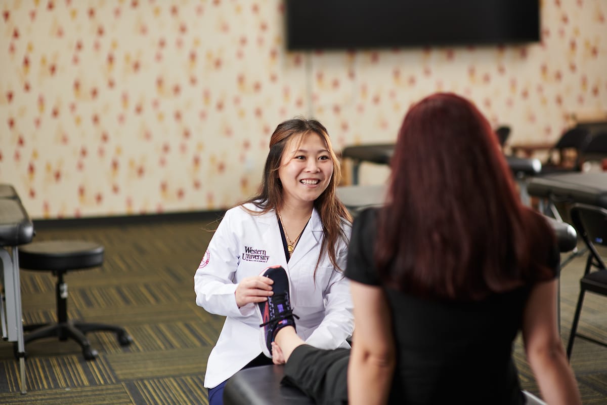 A healthcare professional in a white coat examines a patient's foot in a clinical setting with examination tables and patterned wallpaper.