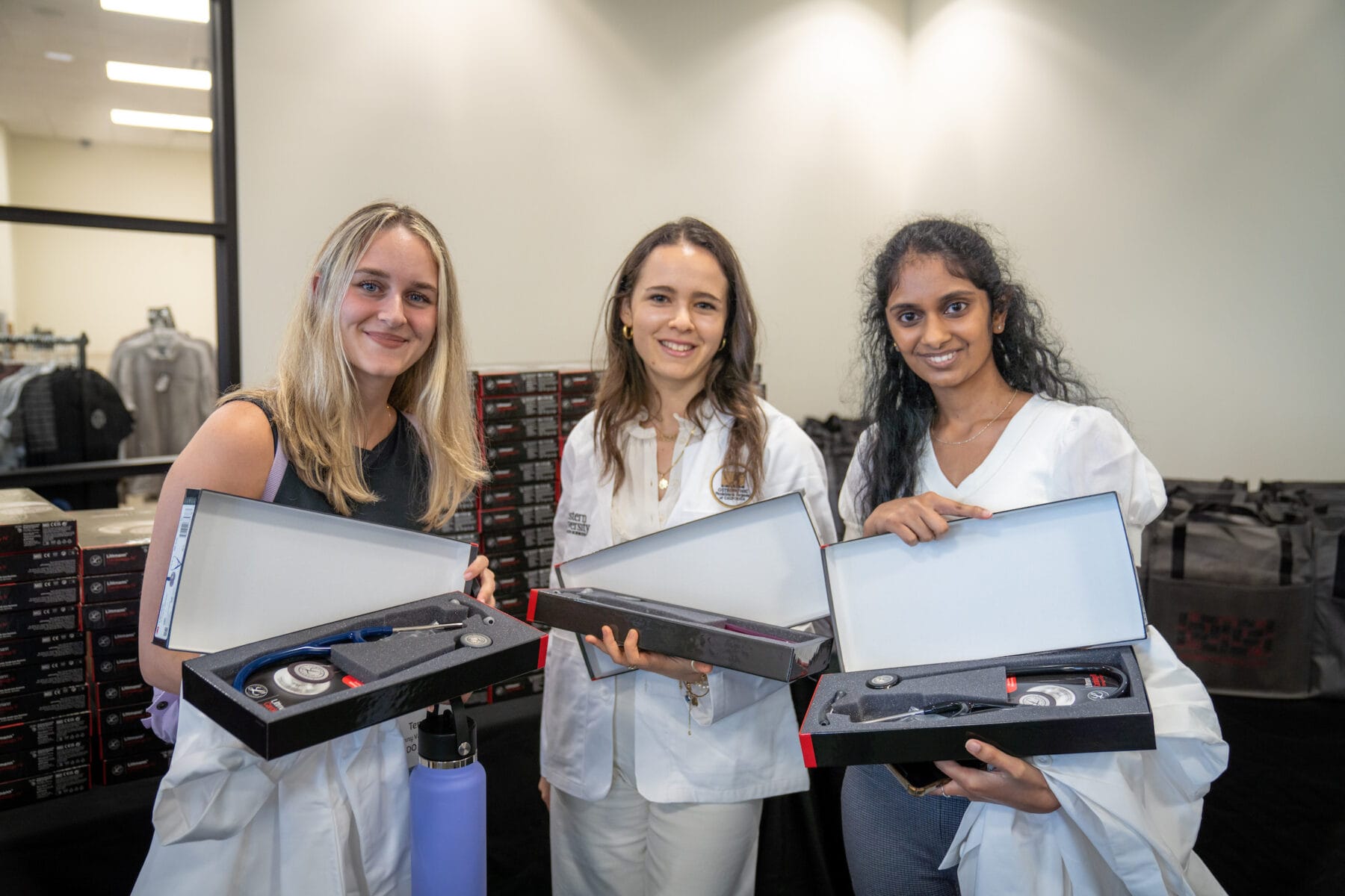 Three women standing indoors, each holding an open box containing a stethoscope and accessories, wearing white coats or medical attire.