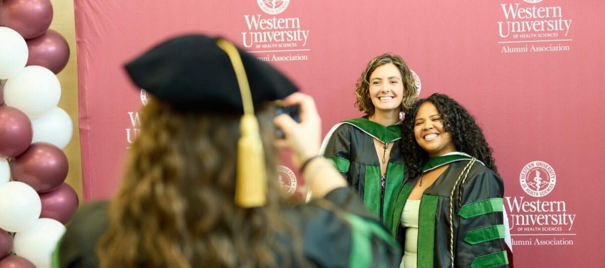 A person is taking a photo of two female college graduates with their hood and gown, smiling and embracing.