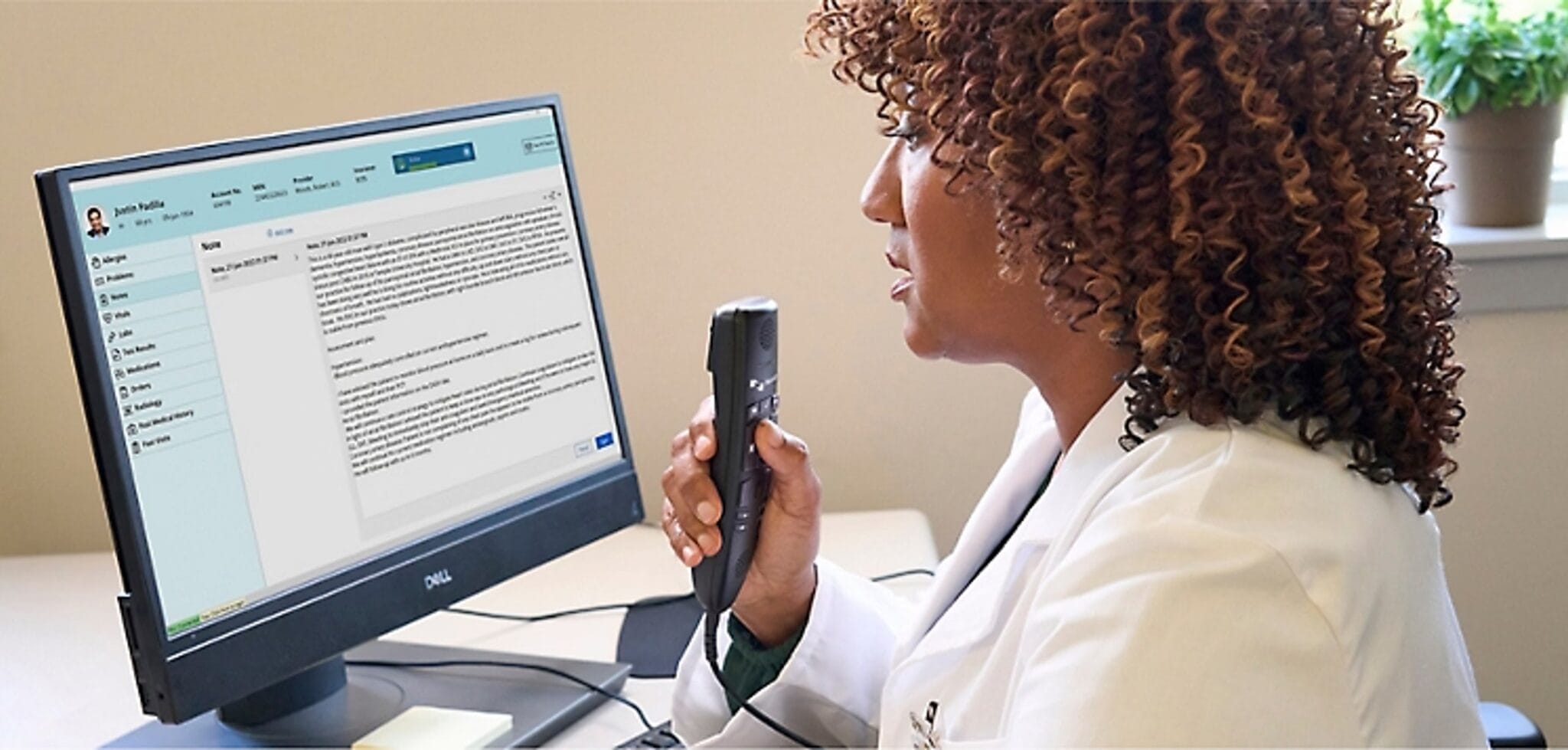 A doctor in a white coat uses a voice recorder to dictate notes into a medical records system displayed on a computer screen.