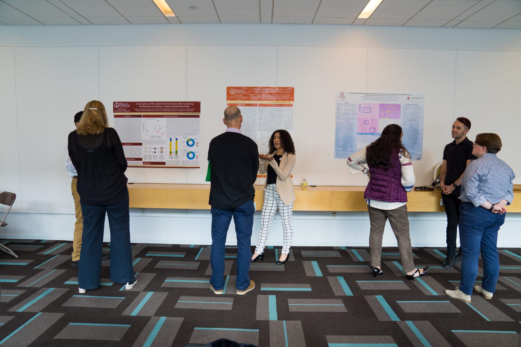 A group of people stand and converse in front of three scientific posters displayed on a wall in a spacious room with patterned carpet.