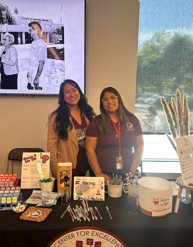 Two people standing behind a table with plants and educational materials, in front of a "WesternU to teach to heal together" sign.