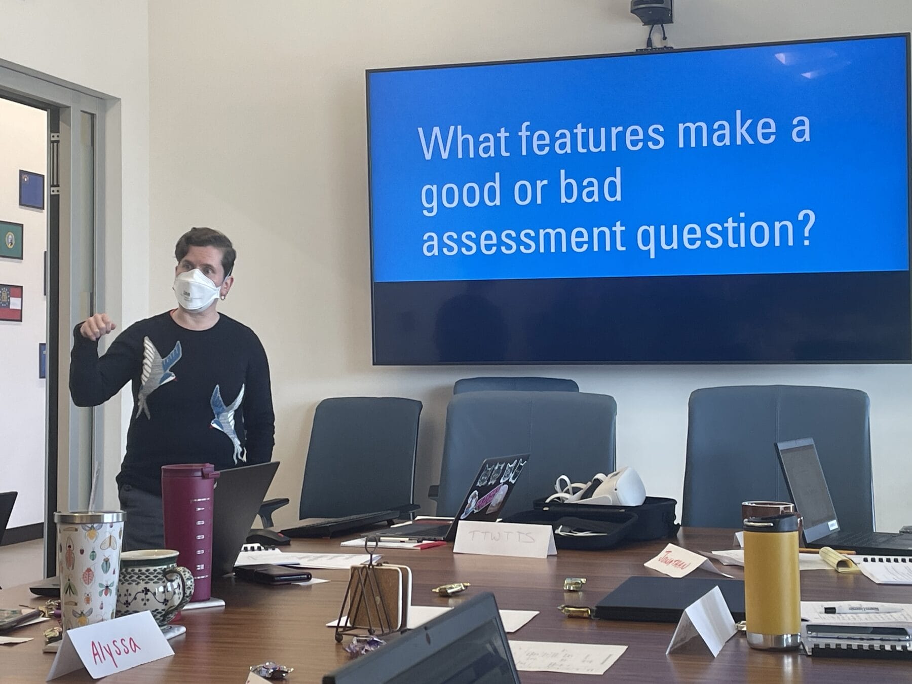 A person wearing a mask presents in a meeting room; a screen behind them displays the text, "What features make a good or bad assessment question?.