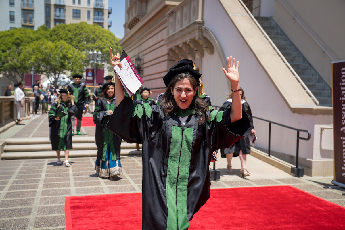 A woman in a graduation gown waving on a red carpet.