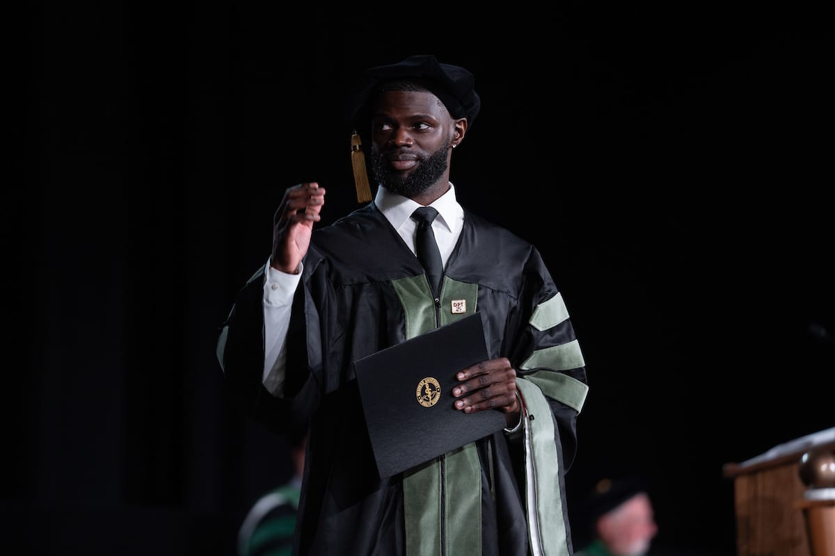 A graduate in a black and green gown holds a diploma, standing on a stage.