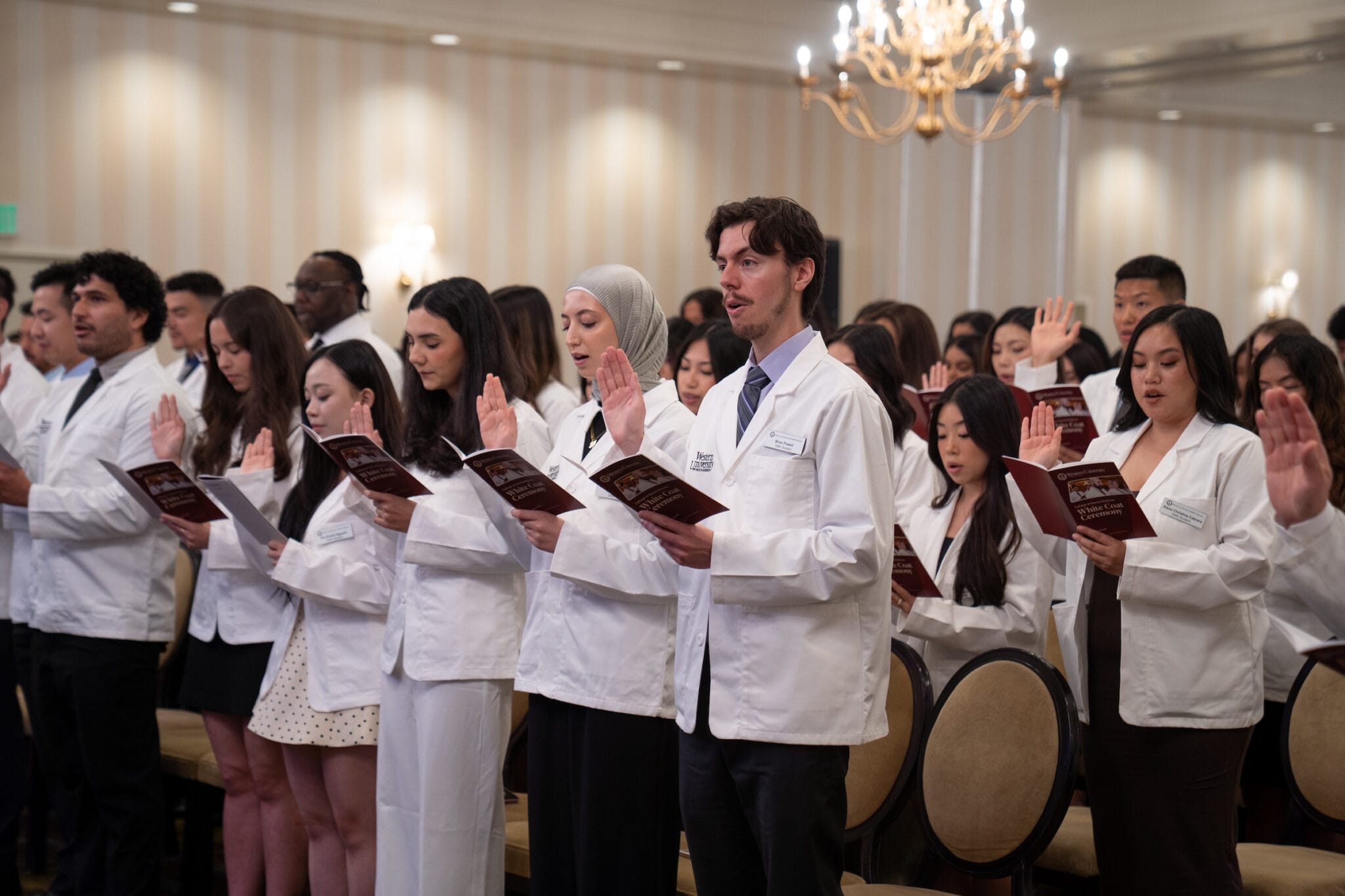 A group of people in white coats stand and raise their right hands while holding booklets, participating in a formal ceremony in a well-lit room.