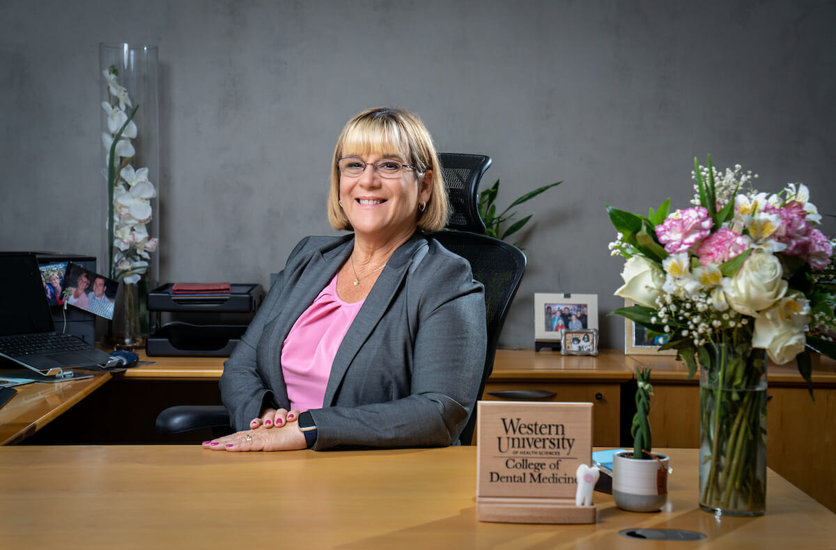 Dental college dean Elizabeth Andrews, wearing gray suit and pink blouse, seated at her desk