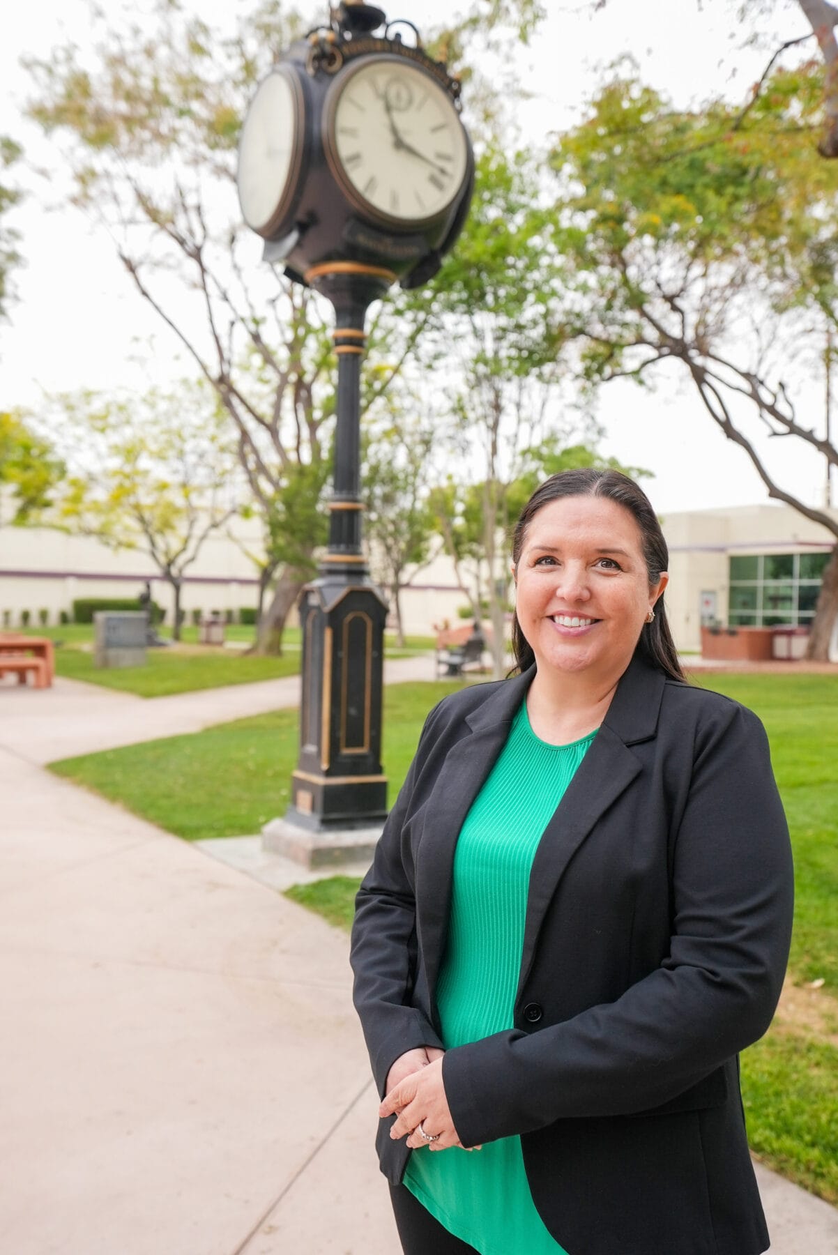 A woman in a green top and black blazer stands smiling in front of an outdoor clock on a college campus walkway. Trees and buildings are visible in the background.