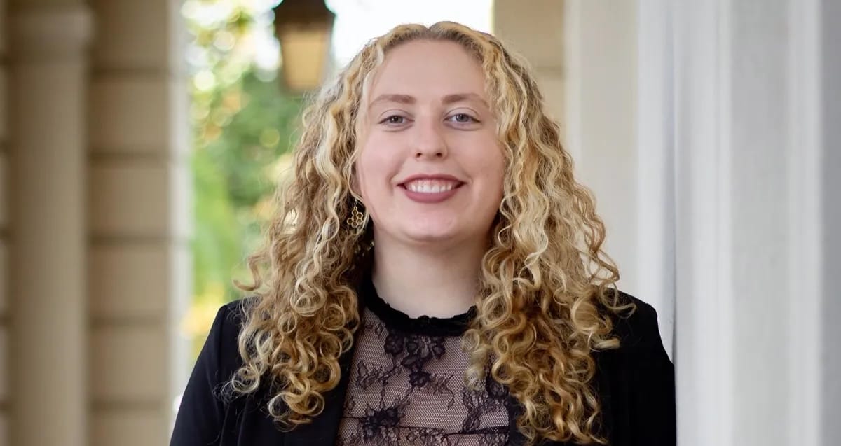 A person with long curly blonde hair, wearing a black lace top and blazer, stands outdoors in front of a building and smiles at the camera.