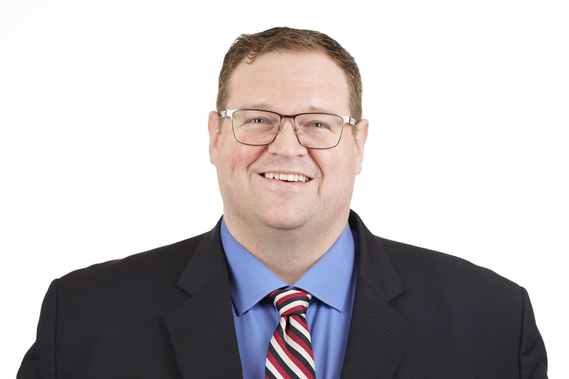Man with short brown hair and glasses, wearing a blue shirt, striped tie, and dark blazer, smiling against a plain white background.
