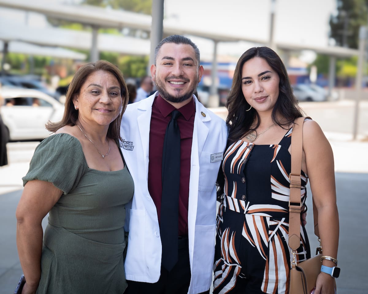 Three people stand together outside, smiling at the camera. The person in the middle is wearing a white coat and tie, and the others are in casual attire.