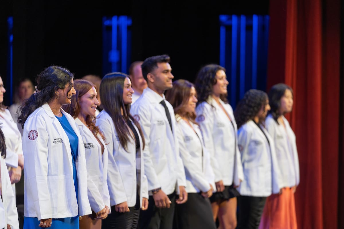 A group of medical students stand in a line on stage, wearing white coats, with red curtains and blue lighting in the background.