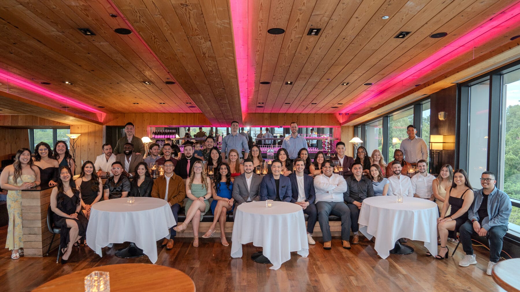 A large group of people pose together at an indoor event space with wooden ceiling and pink lighting, seated and standing around circular tables.