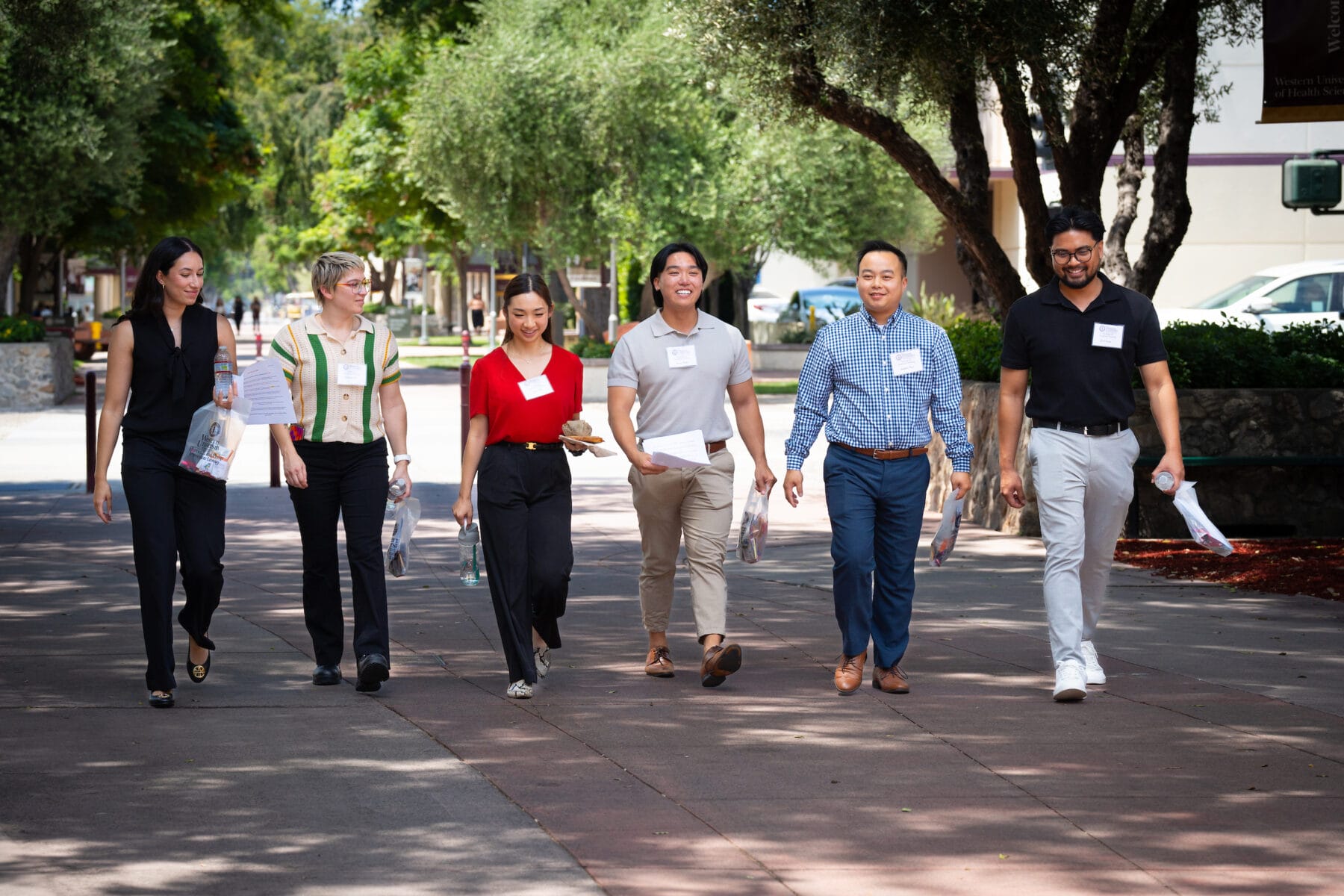 A diverse group of six individuals walks on a sunny outdoor path, each holding papers and wearing name tags. Trees line the sidewalk in the background.