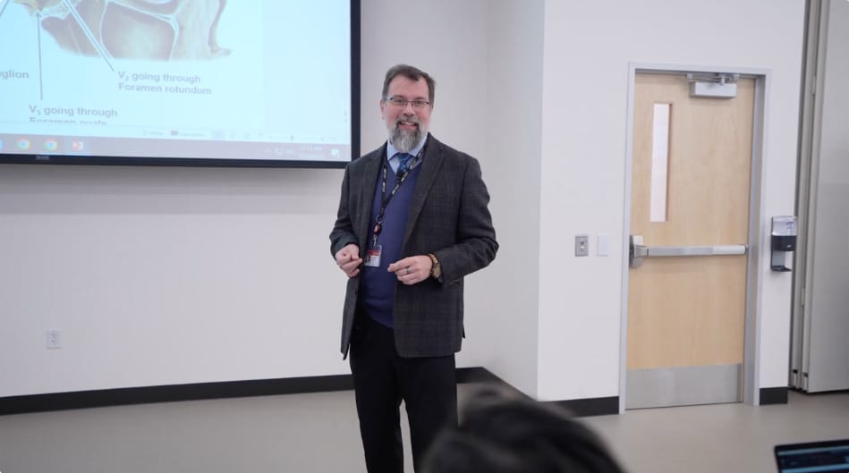 A bearded man in a suit and tie stands in a classroom near a projector screen, giving a presentation. A wooden door is in the background.
