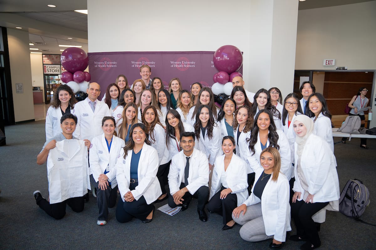 A group of students in lab coats and business attire pose for a photo indoors, with balloons and a banner in the background.
