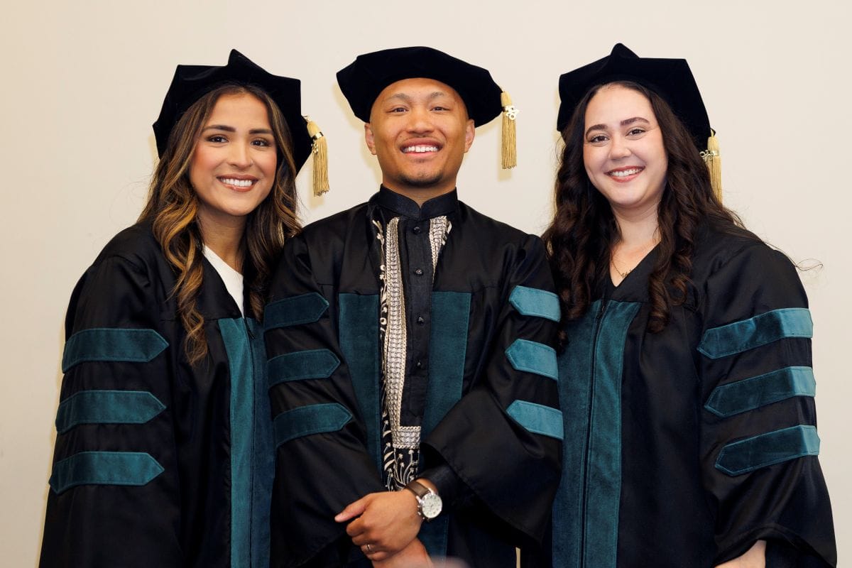 Three graduates in academic regalia stand side by side, smiling at the camera against a plain background.