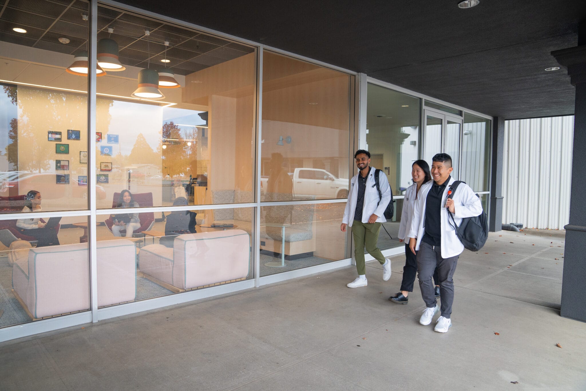 Three people with backpacks walk on a sidewalk outside a building with large windows; several people are visible sitting and talking inside.