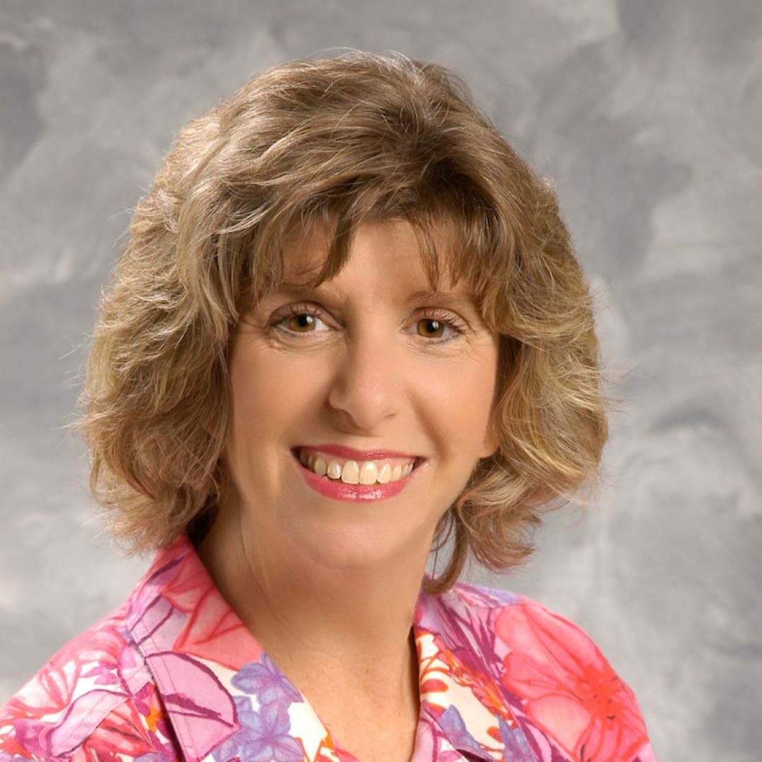 Woman with short, wavy light brown hair smiling at the camera, wearing a pink floral blouse, in front of a gray studio background.