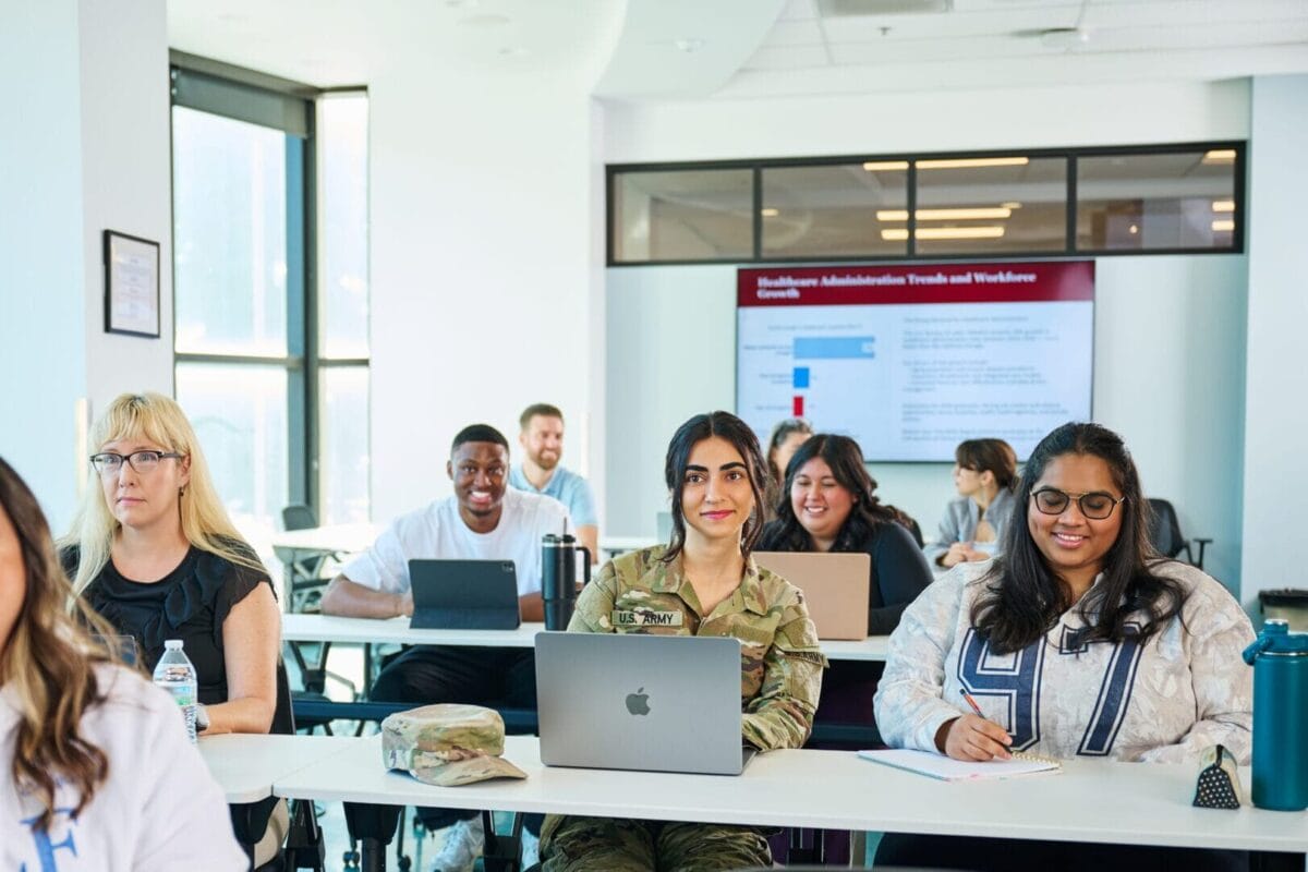 A group of students sit at desks with laptops in a classroom, attentively listening and facing the front.