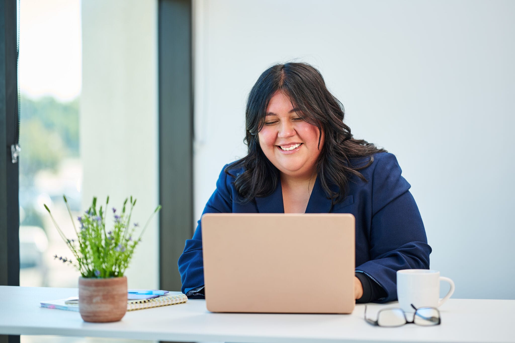 Woman in a navy blazer sits at a desk with a laptop, smiling. A potted plant, notebook, glasses, and coffee mug are also on the desk.