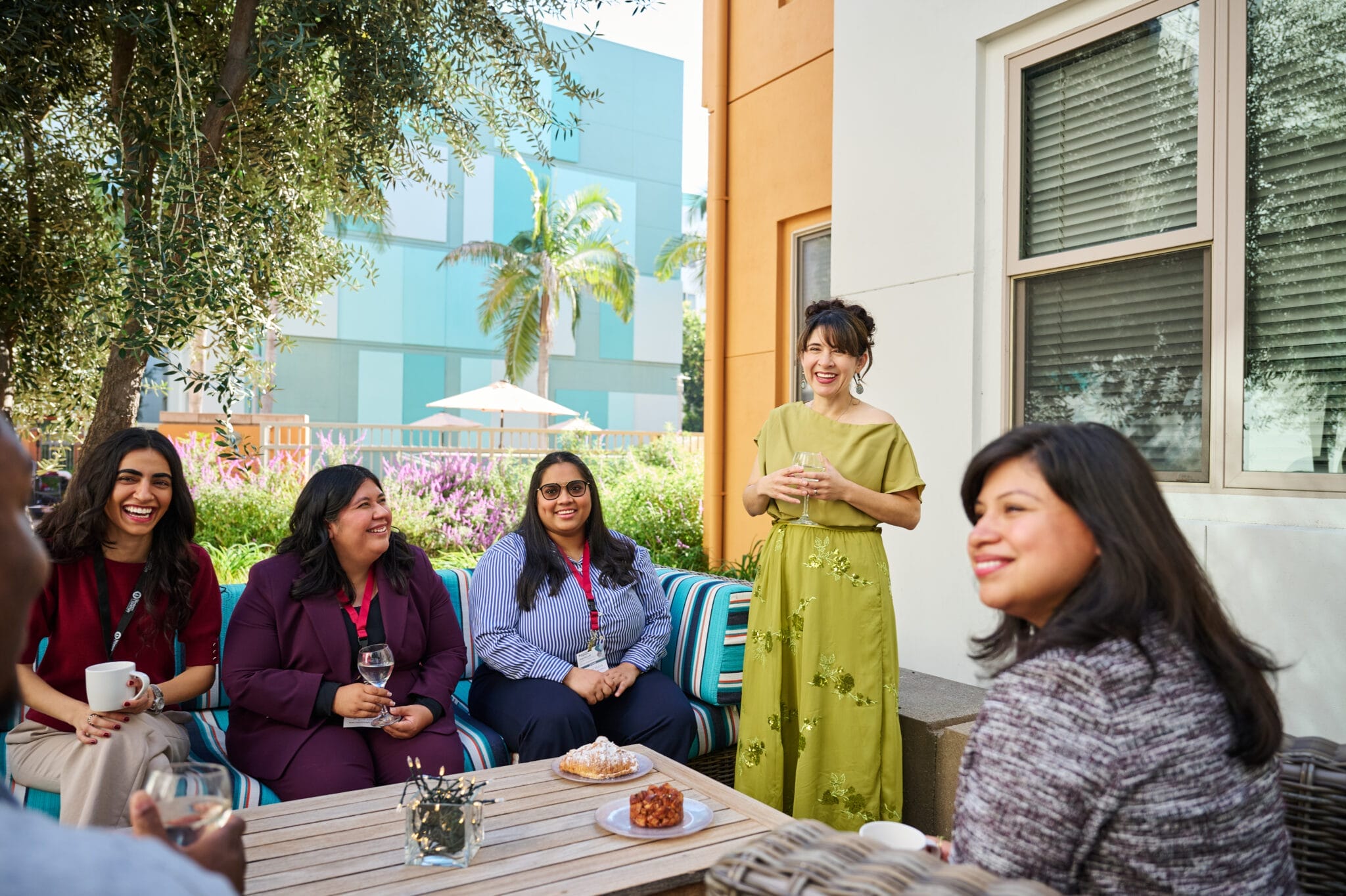 A group of women sit and stand around an outdoor table, talking and smiling, with drinks and snacks in front of them.