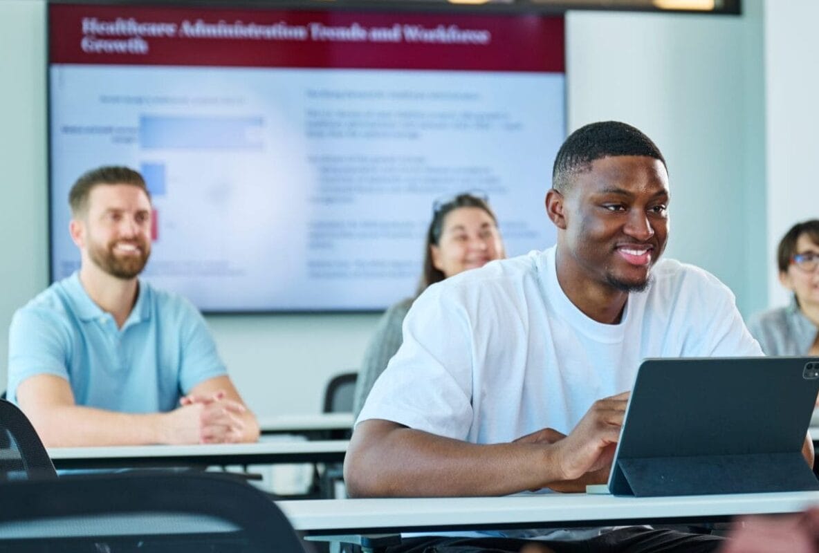 A group of students sits at desks in a classroom, with one student in the foreground using a tablet and a presentation displayed on a screen in the background.