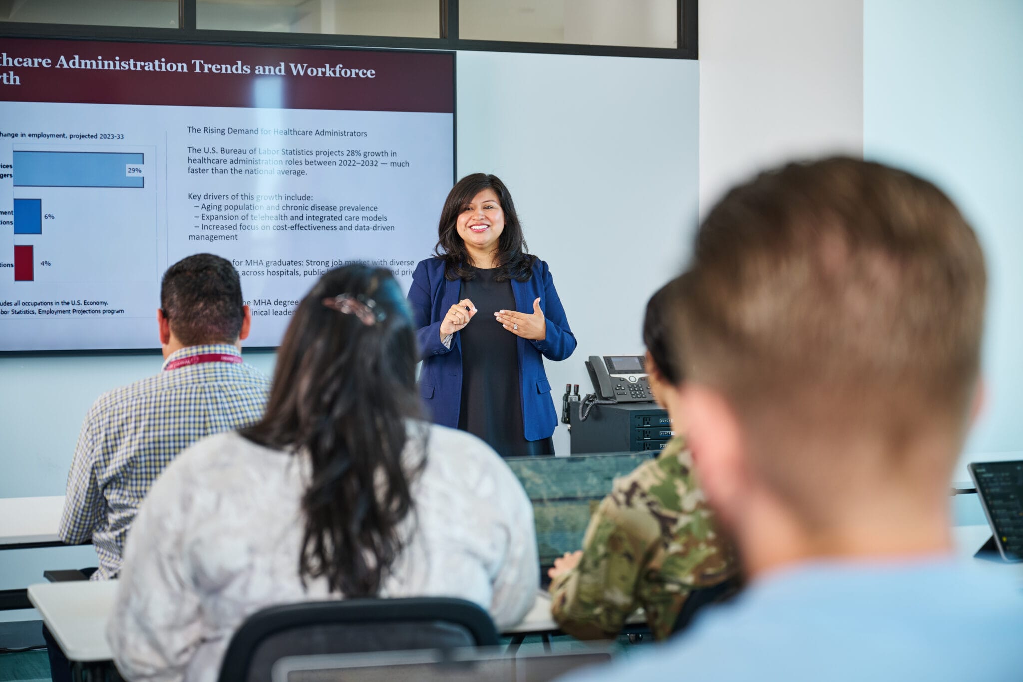 A woman stands in front of a presentation screen discussing healthcare administration trends to a seated audience in a classroom setting.