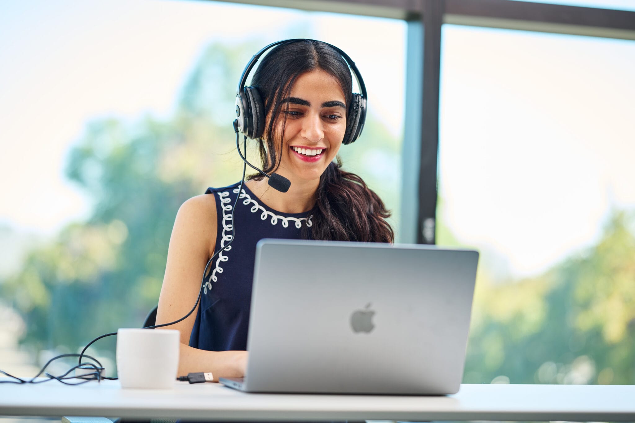 A woman wearing a headset sits at a desk, smiling at her laptop with a coffee mug and USB drive nearby. A large window with trees outside is in the background.