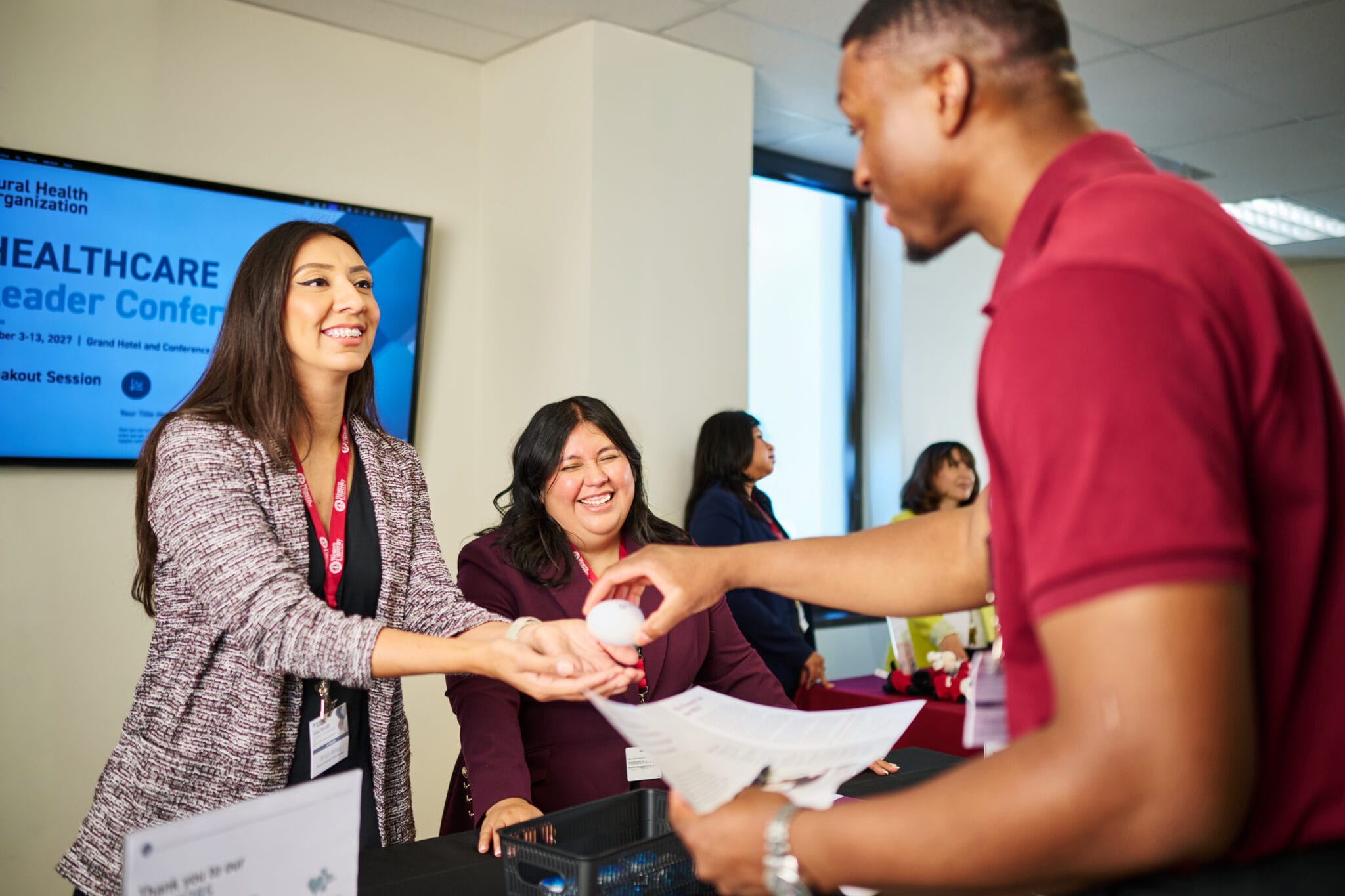 A man hands a small object to a woman at a registration table during a healthcare conference, while another woman smiles in the background.