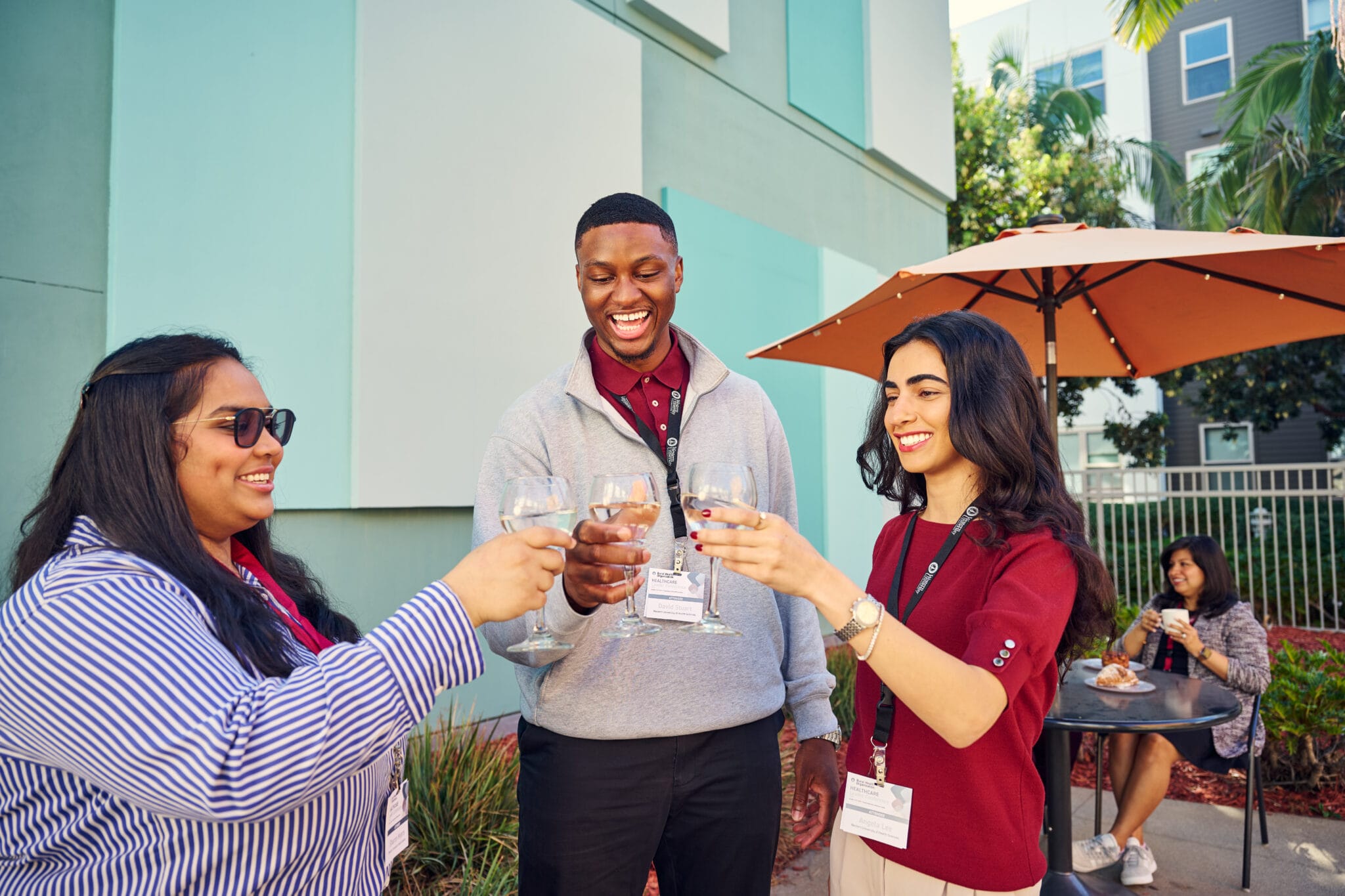 Three people standing outdoors clink glasses and smile, while a fourth person sits at a table in the background near an orange umbrella.