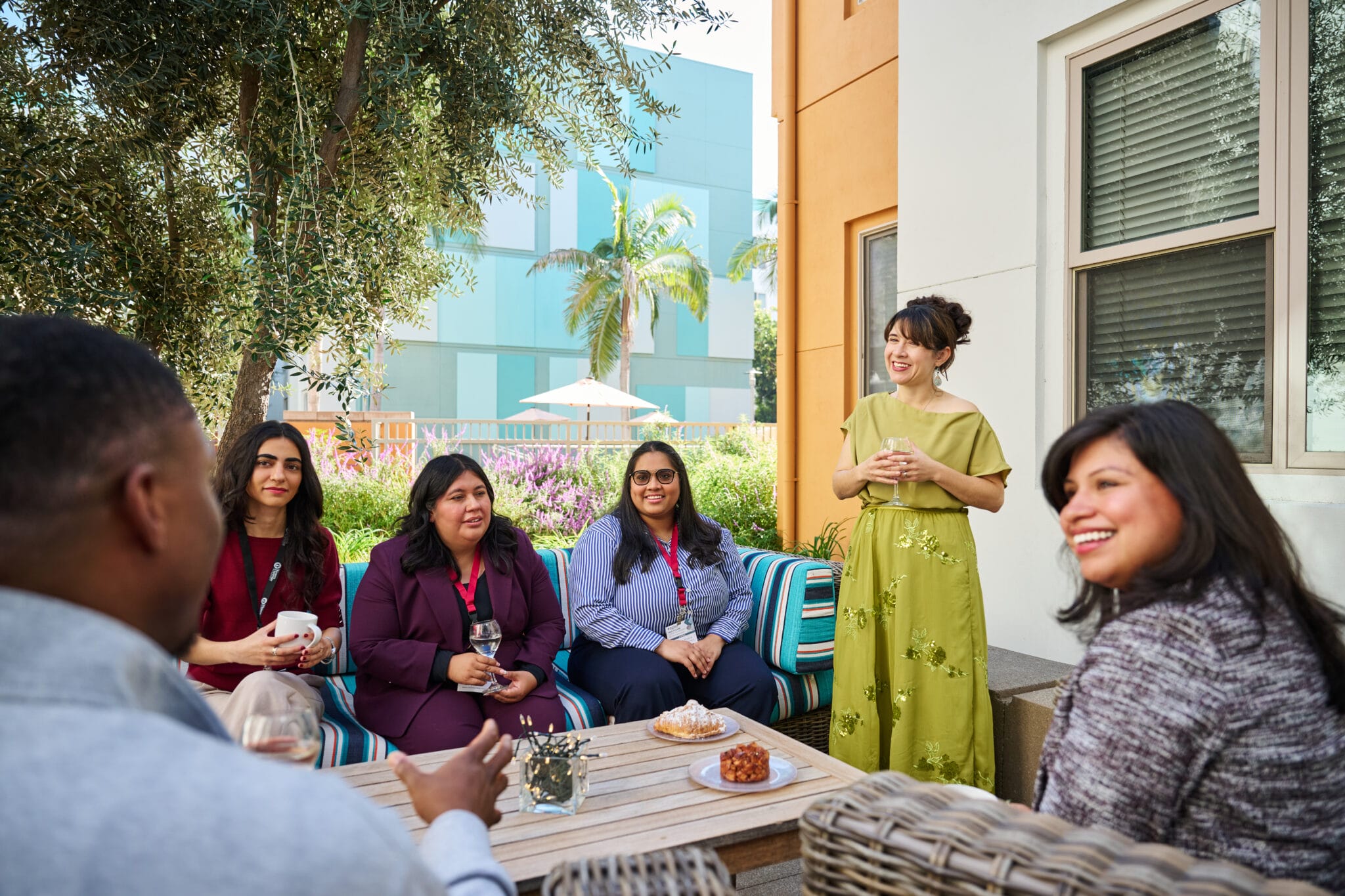 A group of six people sit and stand around an outdoor table, engaged in conversation, with snacks and drinks in front of them.
