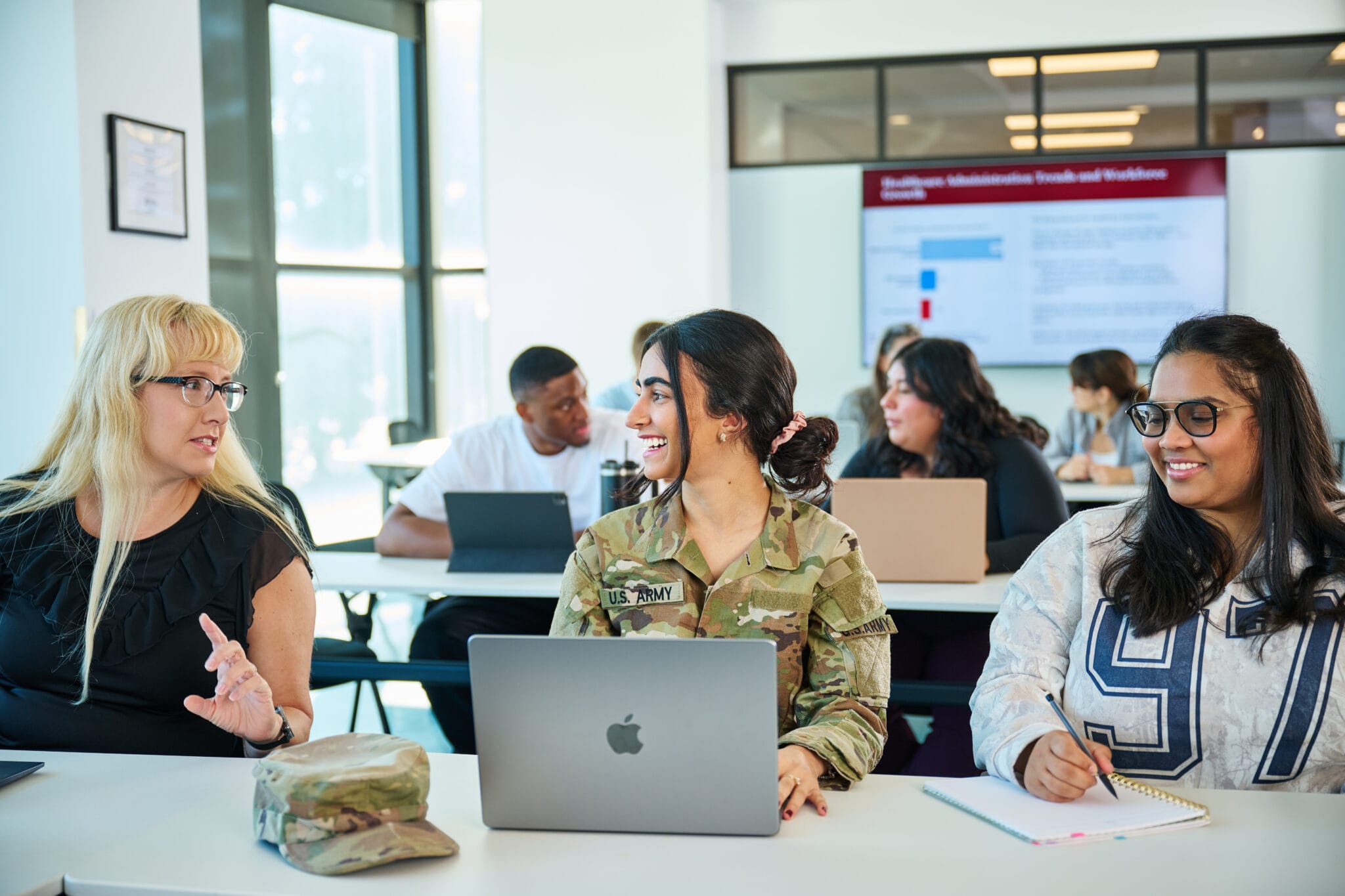 Three women, including one in military uniform, sit at desks with laptops in a classroom, engaged in conversation. Other students and a presentation screen are visible in the background.