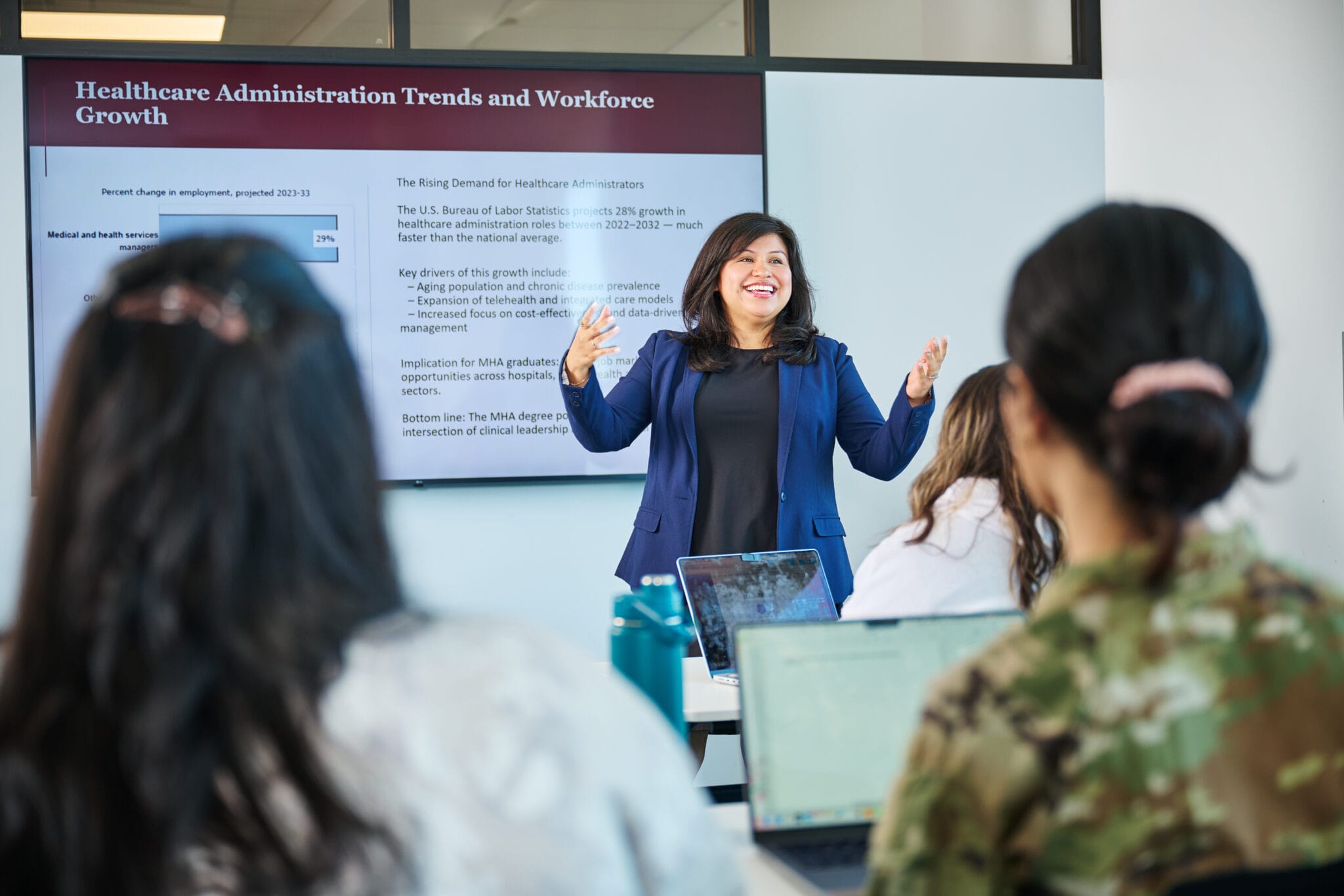 A woman gives a presentation on healthcare administration trends to a group of students in a classroom; a slide is displayed on a screen behind her.