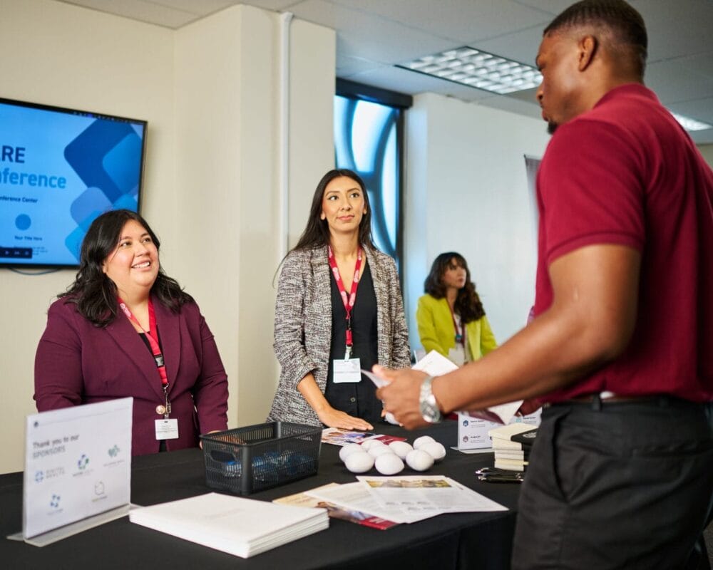 Two women at a conference booth speak with a man. A Healthcare Leader Conference sign is displayed on a screen behind them. Papers, pens, and promotional items are on the table.