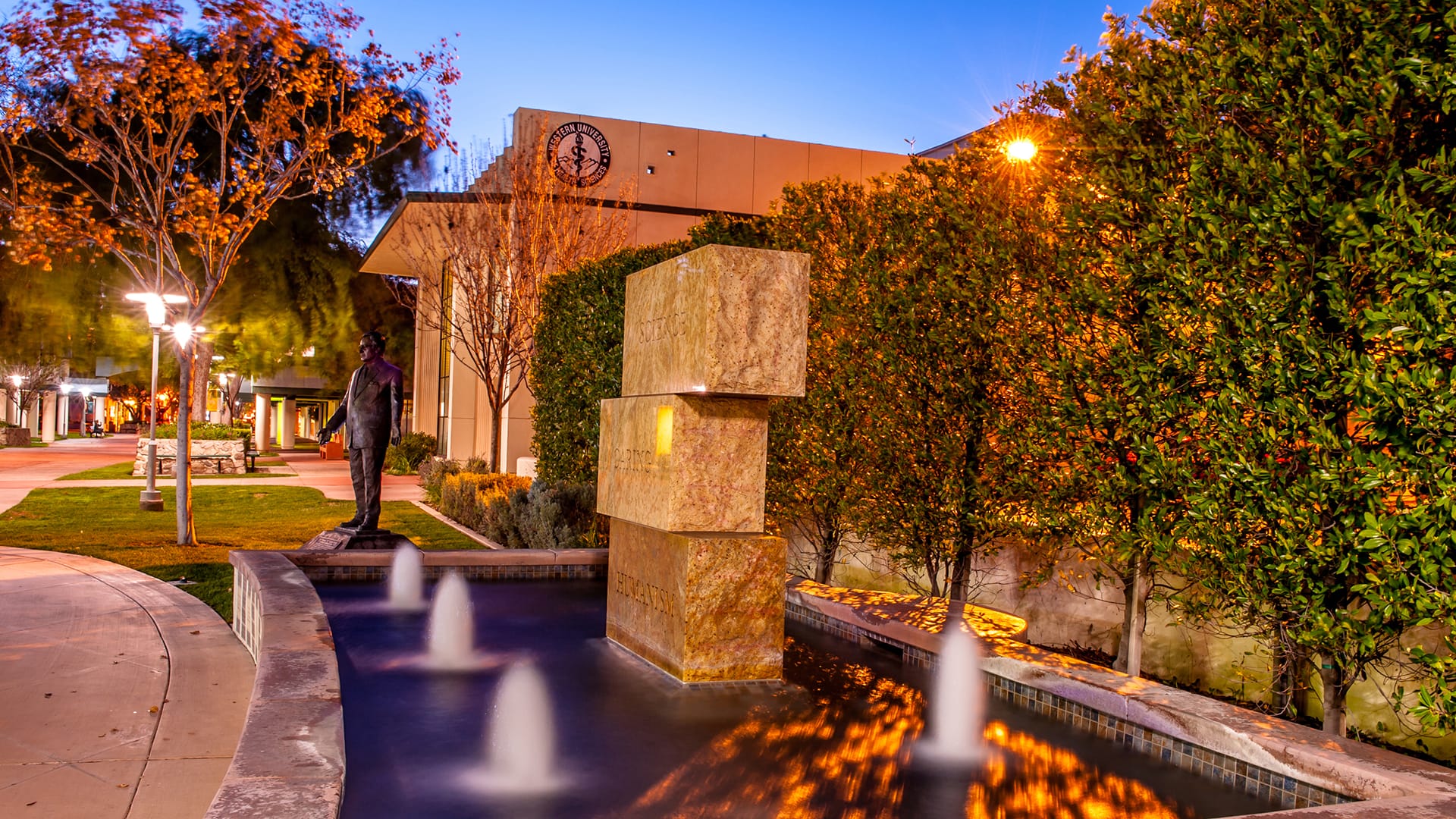 A tiered stone fountain with water jets in front of a building with the seal of Pasadena City College, surrounded by trees and greenery at dusk.