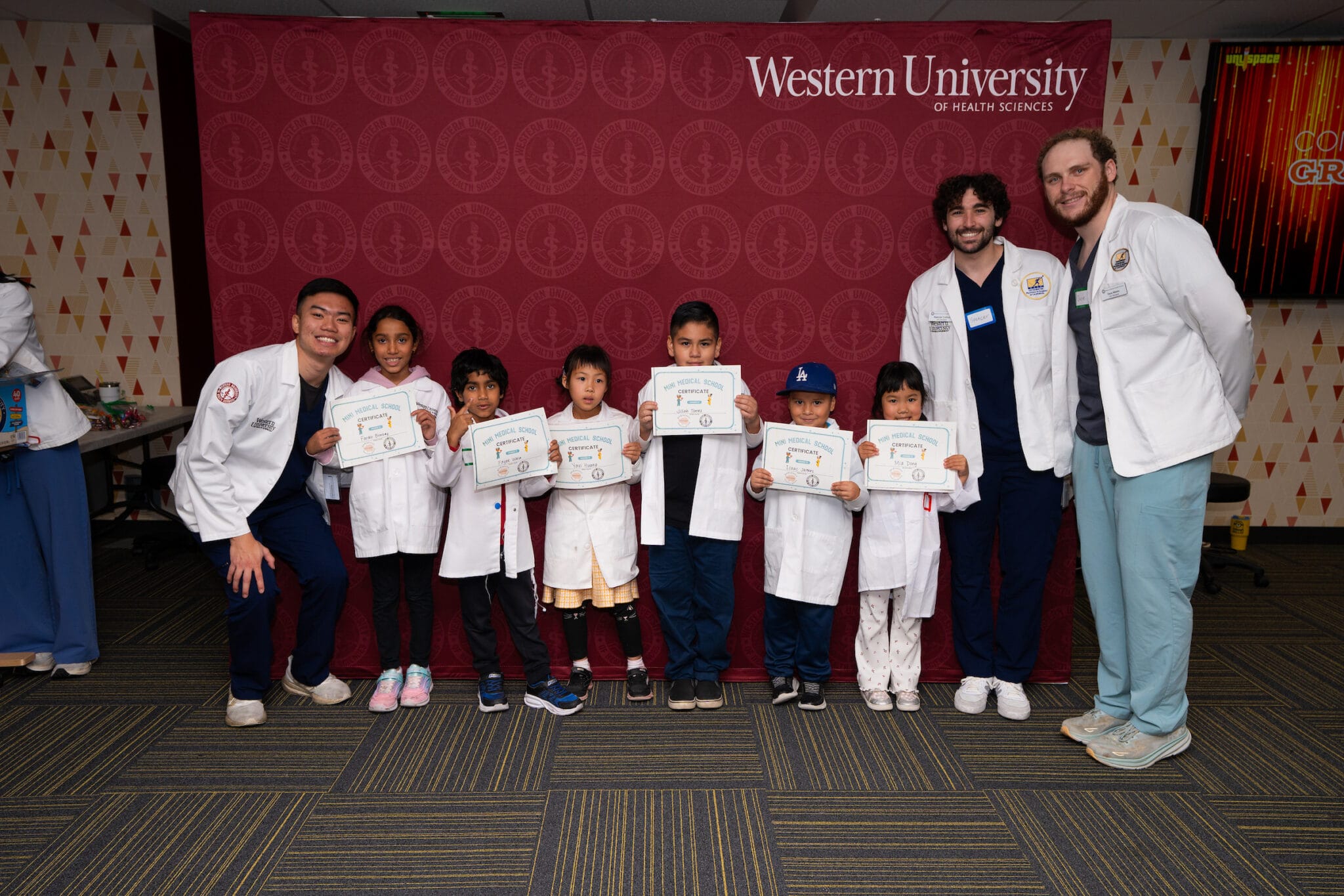 A group of children holding certificates stand in front of a Western University backdrop, flanked by four adults in white coats.