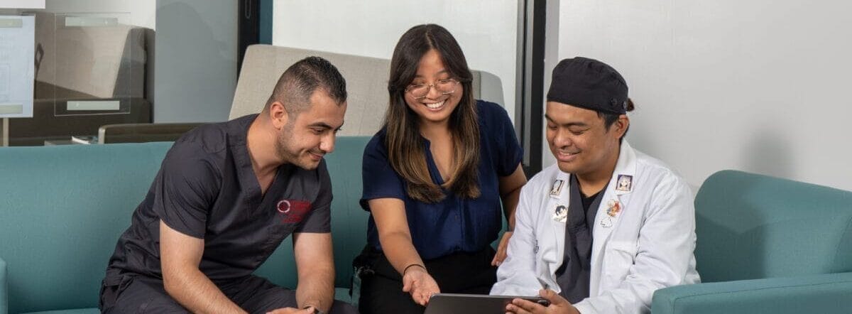 Three people sit on teal couches around a small table, looking at a laptop and tablet in a modern office with glass walls and framed posters.