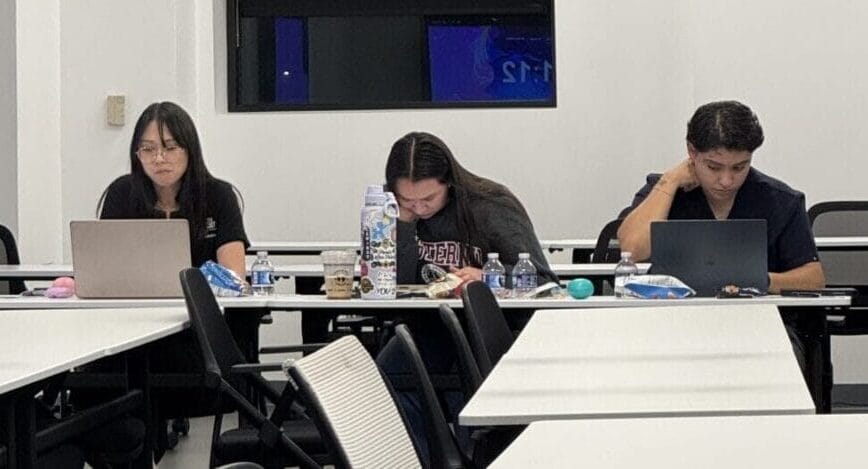 Three people are seated at a table with laptops and papers, working in a mostly empty classroom with rows of unoccupied chairs and desks.